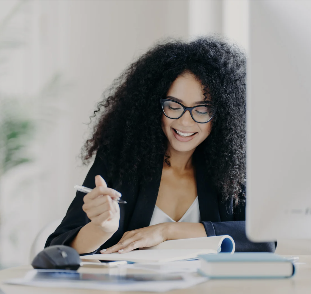 A woman with long hair sits at a desk surrounded by books and technology, smiling while holding a pen and writing.