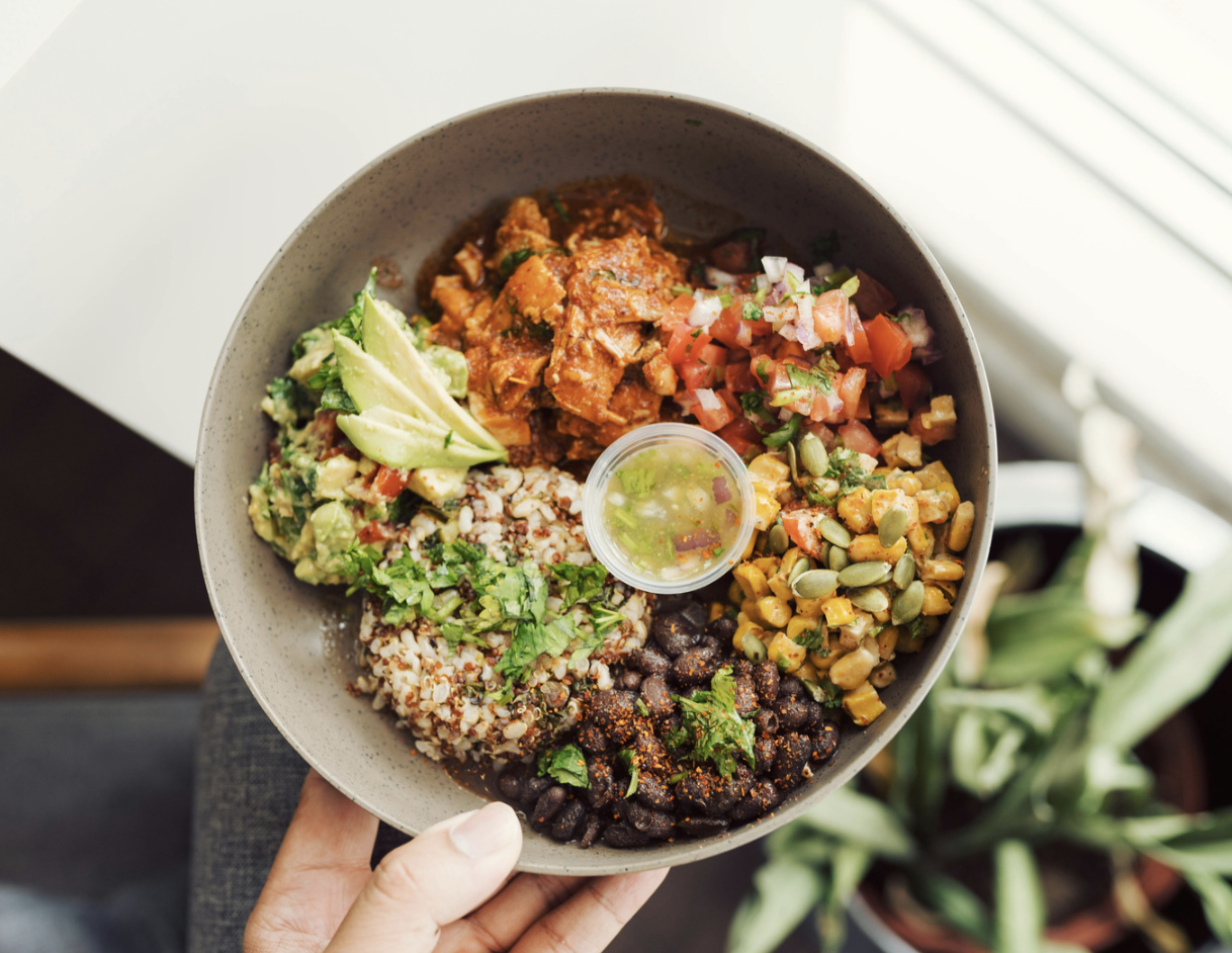 A white bowl filled with a healthy meal, containing a variety of ingredients like avocados, tomatoes, rice, giving an impr...
